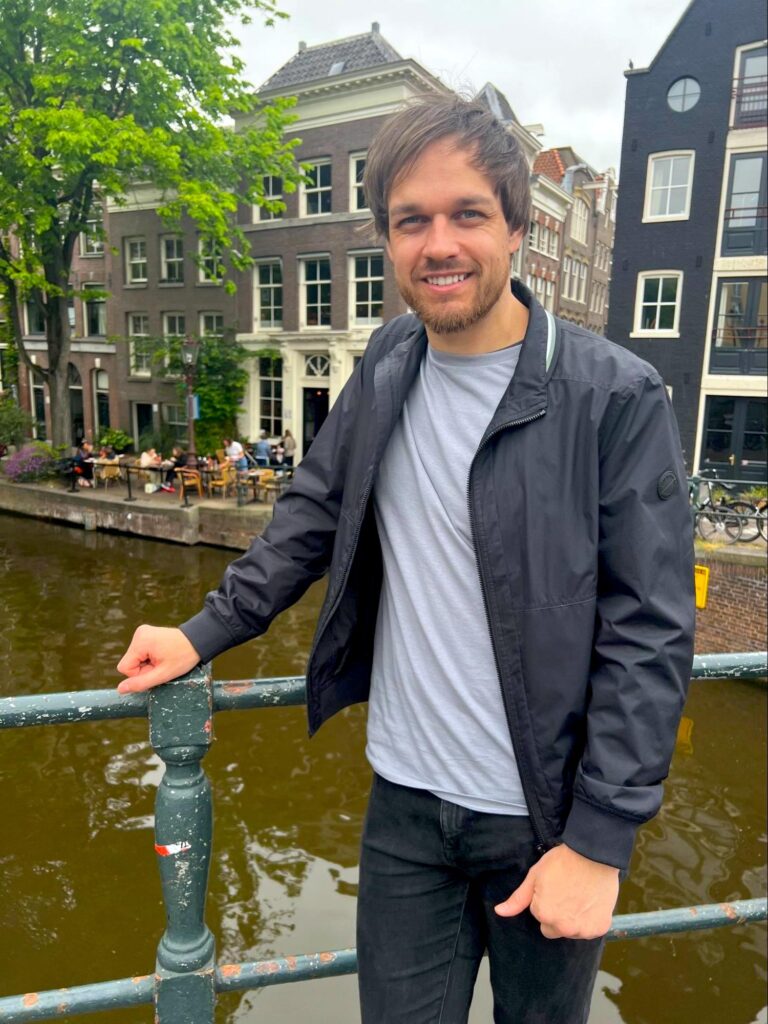 David, a white young man with brown hair, stands on a bridge over an Amsterdam canal.
