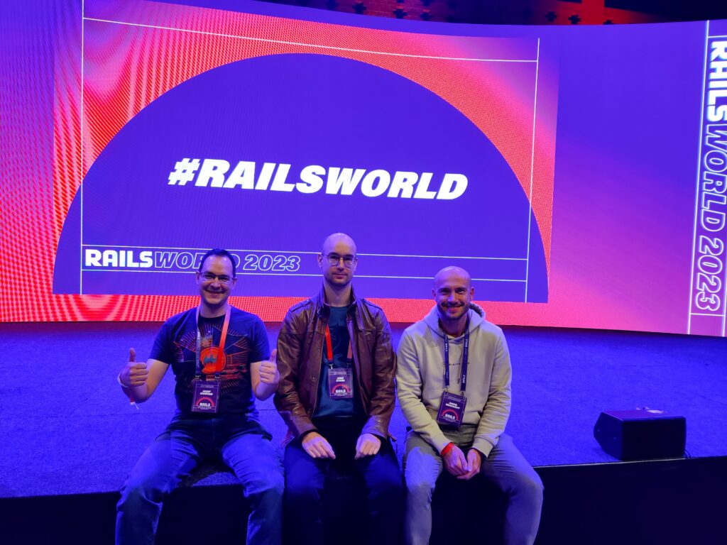 Three primehammer members sitting on the stage of the RailsWorld conference in Amsterdam under a dim blue light, against a screen projecting the Railsworld hashtag and logo in white on blue and pink.