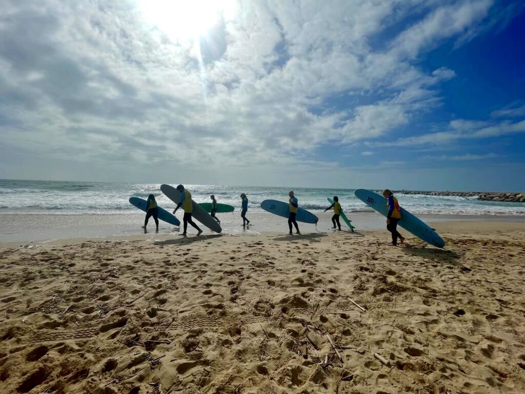 Seven members of the primehammer team walk on a Lisbon beach carrying surf boards under a blue sky.